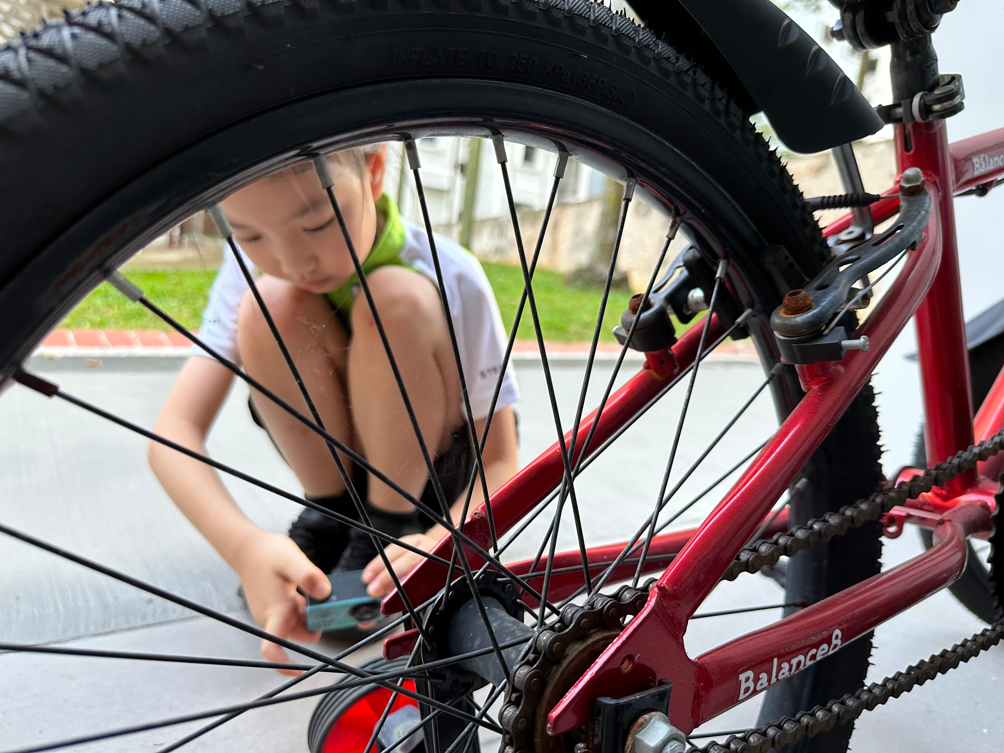 A young child crouches on pavement, working on the rear wheel of a red Balance8 bicycle outdoors.